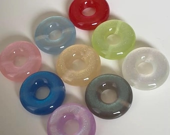 Può includere: Una collezione di nove oggetti circolari a forma di ciambella in vari colori, tra cui rosso, azzurro, rosa, giallo, verde, bianco, blu scuro, grigio e viola. Ogni pezzo presenta una finitura liscia e lucida con sottili particelle glitterate.
