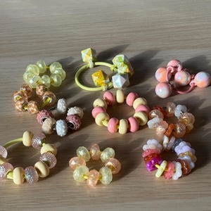 Può includere: Una collezione di elastici per capelli con perline colorate in vari design. Gli elastici per capelli presentano una varietà di perline nelle tonalità del rosa, giallo, arancione e bianco. Alcune perline sono scintillanti, altre geometriche.