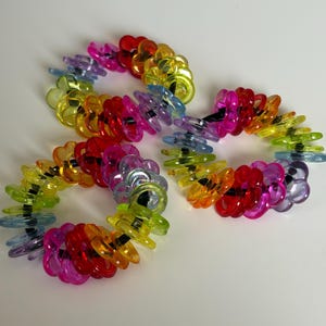 Op de afbeelding: Drie kleurrijke, cirkelvormige haaraccessoires. Elk is samengesteld uit doorschijnende, regenboogkleurige plastic stukken. De kleuren zijn rood, oranje, geel, groen, blauw en paars. De accessoires zijn gerangschikt op een witte ondergrond.