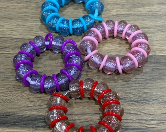 Può includere: Quattro elastici per capelli circolari, ciascuno caratterizzato da una serie di perline rotonde trasparenti con glitter separate da anelli in plastica colorata. Il set include un elastico blu, uno rosa, uno viola e uno rosso con anelli.
