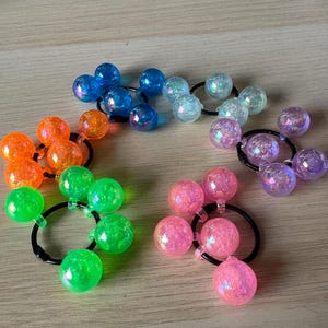 May include: A selection of colourful hair ties, each featuring iridescent, spherical beads. The hair ties are black, and the beads are in shades of blue, orange, green, pink, and purple. The beads have a textured, glittery appearance.