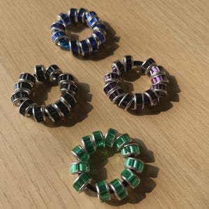 Può includere: Quattro elastici colorati per capelli sono esposti su una superficie di legno. Gli elastici sono circolari, ciascuno ornato con perline rettangolari nei toni del blu, nero, viola e verde. Le perline sono disposte attorno a un elastico nero.