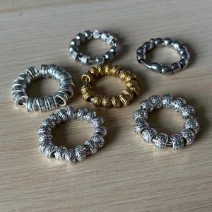 Può includere: Sei anelli metallici, argentati e dorati, con design diversi. Gli anelli presentano varie trame e motivi. Sono disposti su una superficie di legno chiaro, mettendo in risalto i loro dettagli.