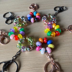 Può includere: Portachiavi con disegni floreali di perline colorate. Ogni portachiavi presenta un anello, un moschettone e una catena, con un fiore fatto di perline di vari colori. Le perline includono perline rosa, blu, gialle, verdi, arancioni e trasparenti.