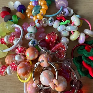 Op de afbeelding: Een verzameling kleurrijke sleutelhangers en accessoires met kralen. De kralen hebben verschillende vormen, maten en kleuren, waaronder rood, oranje, roze, blauw en groen. Sommige sleutelhangers hebben metalen ringen, andere elastiekjes. De afbeelding is van bovenaf genomen.