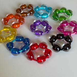 Peut inclure: Plusieurs bracelets fleuris en perles colorées. Les bracelets présentent des perles rondes et transparentes de différentes couleurs : orange, bleu, vert, violet, rose, noir, rouge et jaune. Chaque bracelet est assemblé avec un cordon de couleur assortie.