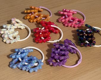 Può includere: Un assortimento di elastici per capelli colorati e perlati in vari colori, tra cui arancione, rosa, rosso, blu, viola e iridescente. Ogni elastico presenta un design circolare con un elastico bianco e un anello colorato coordinato.