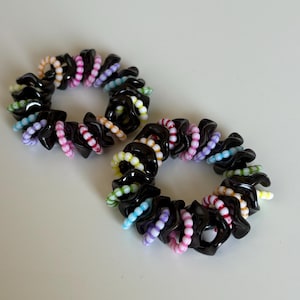 May include: Two black spiral hair ties with colourful beads. Each hair tie has a black, coiled base with a series of small, brightly coloured beads in shades of pink, white, yellow, blue, and green. The hair ties are on a white surface.