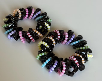 May include: Two black spiral hair ties with colorful beads. Each hair tie has a black, coiled base with a series of small, brightly colored beads in shades of pink, white, yellow, blue, and green. The hair ties are on a white surface.