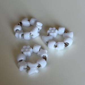May include: Three white beaded rings with heart, flower, and star shapes. The beads are strung on silver-coloured wire, forming a circular design. The rings are displayed on a light-coloured surface.