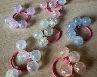 Può includere: Diversi elastici per capelli con perline sferiche iridescenti nei toni del rosa, blu e trasparente. Le perline sono attaccate a elastici rosa. Gli accessori per capelli sono disposti su una superficie di legno chiaro.