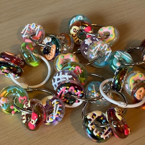 Può includere: Una collezione di portachiavi con perline di vetro colorate. Le perline hanno la forma di ciambelle con vari condimenti e colori, tra cui rosa, bianco e cioccolato. Ogni portachiavi ha un anello di metallo argentato.