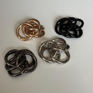 May include: Four metal clasp connectors in gold, black, silver, and gunmetal colours. Each connector is made of interlocking circular rings. The clasps are arranged on a white surface.
