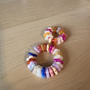 May include: Two colourful beaded bracelets. The bracelets are made of small, flat, irregularly shaped beads in shades of pink, orange, white, blue, and tan. The bracelets are arranged on a light brown wooden surface.
