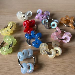 May include: A collection of colorful, beaded keychains. The keychains feature various colors, including yellow, red, blue, pink, and iridescent beads. Each keychain has a silver-colored ring. The beads are round and smooth.
