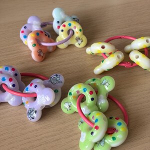 Op de afbeelding: Een verzameling kleurrijke babybijtringen in verschillende pastelkleuren, waaronder geel, groen en paars. Elke bijtring heeft een uniek ontwerp met een ring en kleine, decoratieve vormen. De bijtringen zijn verbonden door roze ringen.