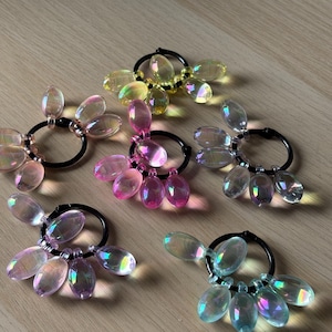 Può includere: Cinque elastici per capelli iridescenti con anelli circolari neri e perline a goccia in vari colori, tra cui rosa, giallo e blu. Le perline hanno un effetto cangiante. Gli accessori per capelli sono disposti su una superficie di legno marrone chiaro.