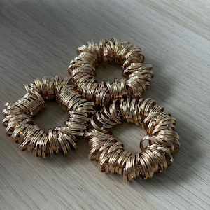 May include: Three gold-coloured hair ties, each made of numerous small, coiled metal rings. The hair ties are arranged on a light-coloured wooden surface, with soft shadows adding depth. The overall aesthetic is simple and elegant.