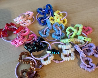 Può includere: Una collezione di elastici colorati per capelli in varie tonalità, tra cui rosa, blu, giallo e viola. Gli elastici presentano disegni decorativi a forma di cuore e geometrici. Gli elastici sono di colore rosa chiaro.