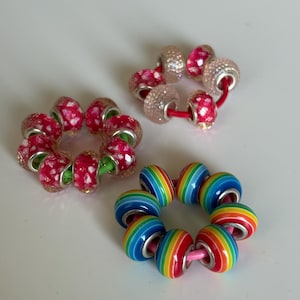 Peut inclure: Trois ensembles de perles décoratives. Deux ensembles présentent des motifs floraux rouges et blancs, enfilés sur des cordons verts et rouges. Le troisième ensemble a un motif rayé arc-en-ciel, enfilé sur un cordon rose. Ces perles sont adaptées à la fabrication de bijoux.