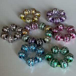 Pode incluir: Uma coleção de pulseiras de contas coloridas. Cada pulseira apresenta um anel de contas decorativas em várias cores, incluindo prata, ouro, roxo, rosa, verde e azul. As contas têm um design padronizado e são enfiadas em bandas elásticas.