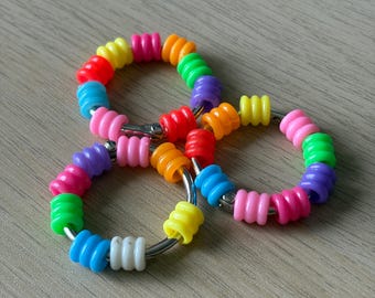 Può includere: Un giocattolo astratto e colorato composto da tre anelli interconnessi. Ogni anello è composto da vivaci perline a spirale in un arcobaleno di colori, tra cui rosso, arancione, giallo, verde, blu e rosa. Le perline sono infilate su anelli di metallo color argento.