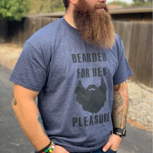 Peut inclure: T-shirt bleu marine avec l'inscription noire "BEARDED FOR HER PLEASURE". Le t-shirt présente un dessin de barbe noire. La personne porte un jean bleu et une montre noire.