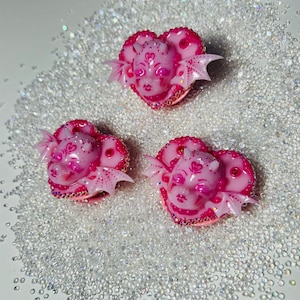 May include: Three pink heart-shaped resin charms with a detailed sculpted face of a creature with wings and horns. The charms are decorated with glitter and rhinestones.