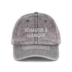 May include: Gray baseball cap with the text "SCHAEFER & LUSAQUE" in white. The cap has a curved brim and a button on top. The fabric has a faded, vintage look.