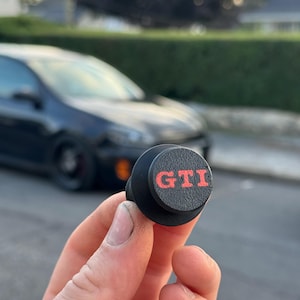 Puede incluir: Una pieza de automóvil negra con las letras rojas "GTI" en una superficie circular. La pieza se sostiene en una mano, con un fondo borroso de un coche y una calle residencial.
