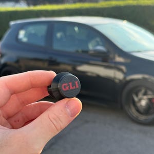 以下が含まれることがあります： 黒と赤のGLIエンブレムが手に持たれており、背景には黒い車があります。エンブレムは円形で、GLIの文字が赤色で表示されています。車は黒色で、舗装された路面に駐車されています。