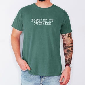 May include: A green t-shirt with the text "POWERED BY GUINNESS" in white block letters. The shirt is a solid color and has short sleeves. The person wearing the shirt is also wearing blue jeans.
