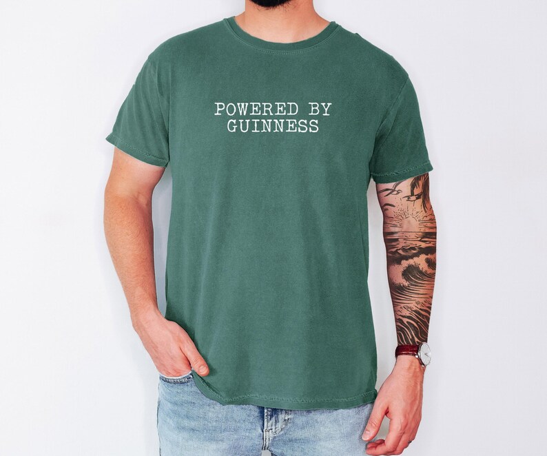 May include: A green t-shirt with the text "POWERED BY GUINNESS" in white block letters. The shirt is a solid color and has short sleeves. The person wearing the shirt is also wearing blue jeans.