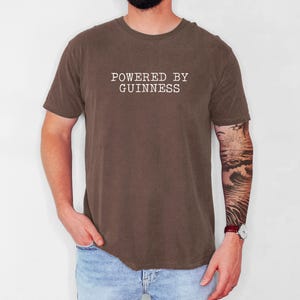 May include: A brown t-shirt with the text "POWERED BY GUINNESS" in white, centered on the chest. The shirt is short-sleeved and has a crew neck. The person is wearing blue jeans.
