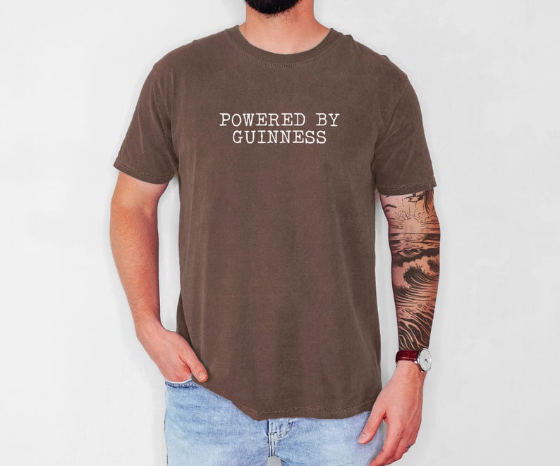May include: A brown t-shirt with the text "POWERED BY GUINNESS" in white, centered on the chest. The shirt is short-sleeved and has a crew neck. The person is wearing blue jeans.