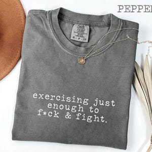 May include: A gray Comfort Colors t-shirt with the text "exercising just enough to f*ck & fight" in white. A silver necklace with a pendant and a brown hat are also visible.