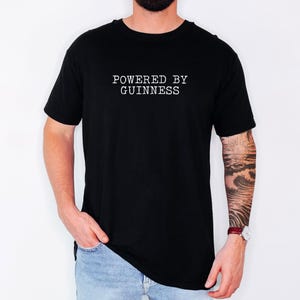 May include: Black t-shirt with the text "POWERED BY GUINNESS" in white, centered on the chest. The shirt is short-sleeved and worn by a person in light blue jeans. The person has a tattoo on their arm and a watch.