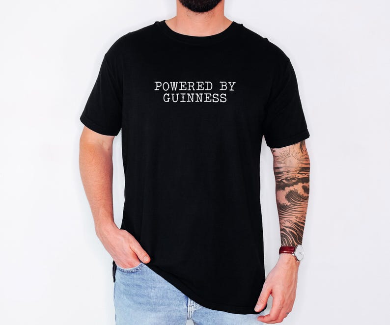 May include: Black t-shirt with the text "POWERED BY GUINNESS" in white, centered on the chest. The shirt is short-sleeved and worn by a person in light blue jeans. The person has a tattoo on their arm and a watch.