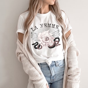 May include: White t-shirt with a graphic design featuring a rose with wings and a snake. The text "LA FEMME" is above the rose.