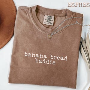 May include: Espresso-coloured t-shirt with the text "banana bread baddie" in white. The shirt has a crew neck and a small tag. A silver necklace with a pendant is draped across the shirt. A brown hat and dried plant are in the background.