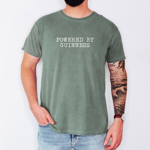 May include: A sage green t-shirt with the text "POWERED BY GUINNESS" printed in white. The shirt is made of a soft material and has a classic crew neck design. The model is wearing blue jeans and a watch.