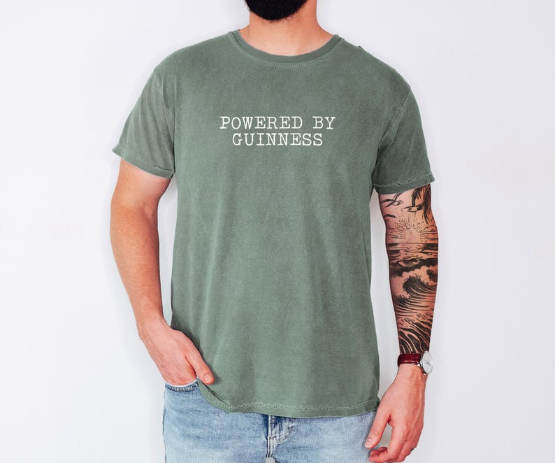 May include: A sage green t-shirt with the text "POWERED BY GUINNESS" printed in white. The shirt is made of a soft material and has a classic crew neck design. The model is wearing blue jeans and a watch.