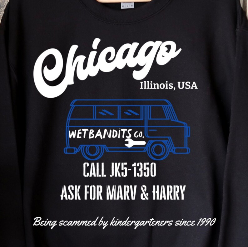 May include: Black sweatshirt with white and blue graphic text. The text reads "Chicago, Illinois, USA" above a blue van illustration. Below the van are the words "WETBANDITS CO." and a phone number. The phrase "Being scammed by kindergarteners since 1990" is at the bottom.