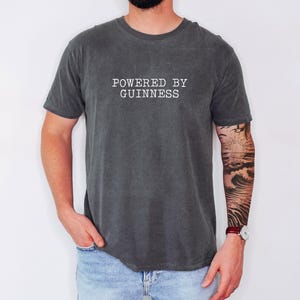 May include: Dark gray t-shirt with the text "POWERED BY GUINNESS" in white. The shirt is a crew neck style and has short sleeves. The person wearing the shirt has a tattoo on their arm.