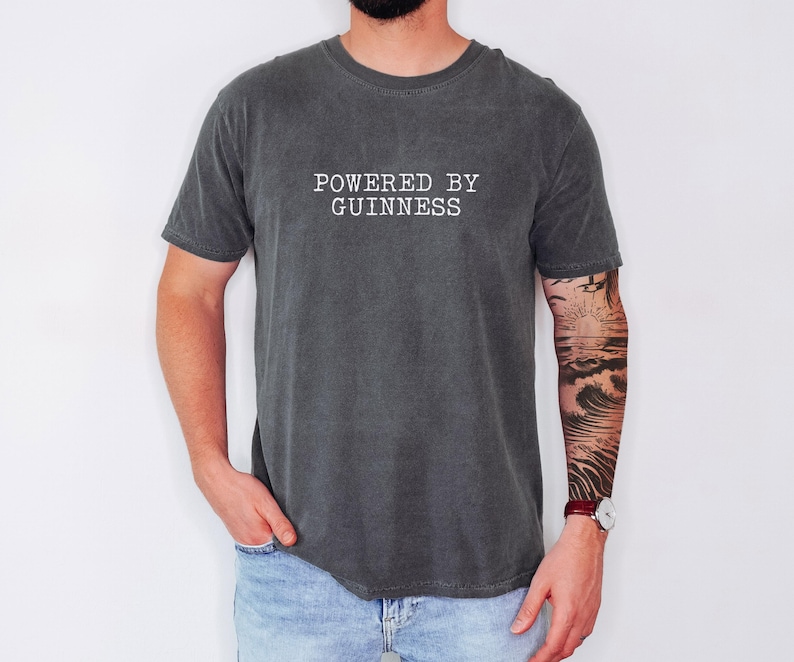 May include: Dark gray t-shirt with the text "POWERED BY GUINNESS" in white. The shirt is a crew neck style and has short sleeves. The person wearing the shirt has a tattoo on their arm.