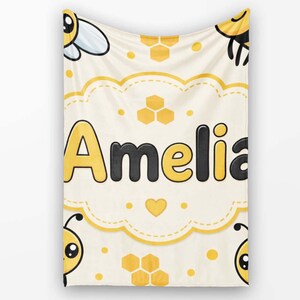May include: A cream-coloured blanket featuring a repeating pattern of cartoon bees and yellow honeycomb shapes. In the centre, the name Amelia is printed in large, bold, black and yellow letters inside a scalloped, yellow-bordered frame.