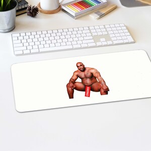May include: A white desk mat featuring a seated, muscular man with a red "CENSORED" banner covering his groin. The man is bald and has a dark complexion. The mat is on a white desk with a keyboard and other office supplies.