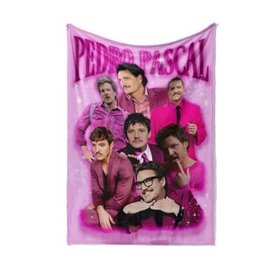 May include: A pink blanket with a collage of images of Pedro Pascal. The name "PEDRO PASCAL" is written in large, pink, bubble letters at the top. The images show Pascal in various poses and outfits, all in shades of pink and purple.