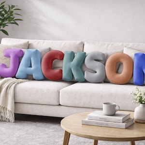 May include: A sofa displays the name "JACKSON" spelled out with plush, colourful letter cushions. The letters are purple, light blue, red, teal, grey, orange, and blue. A beige throw blanket and decorative cushions complete the scene.