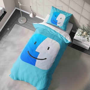 May include: A light blue bed set with a large, stylized face design in white and blue. The set includes a duvet cover and a pillowcase, both with the same graphic. The bedding is on a white bed, with a gray rug and a lamp in the background.